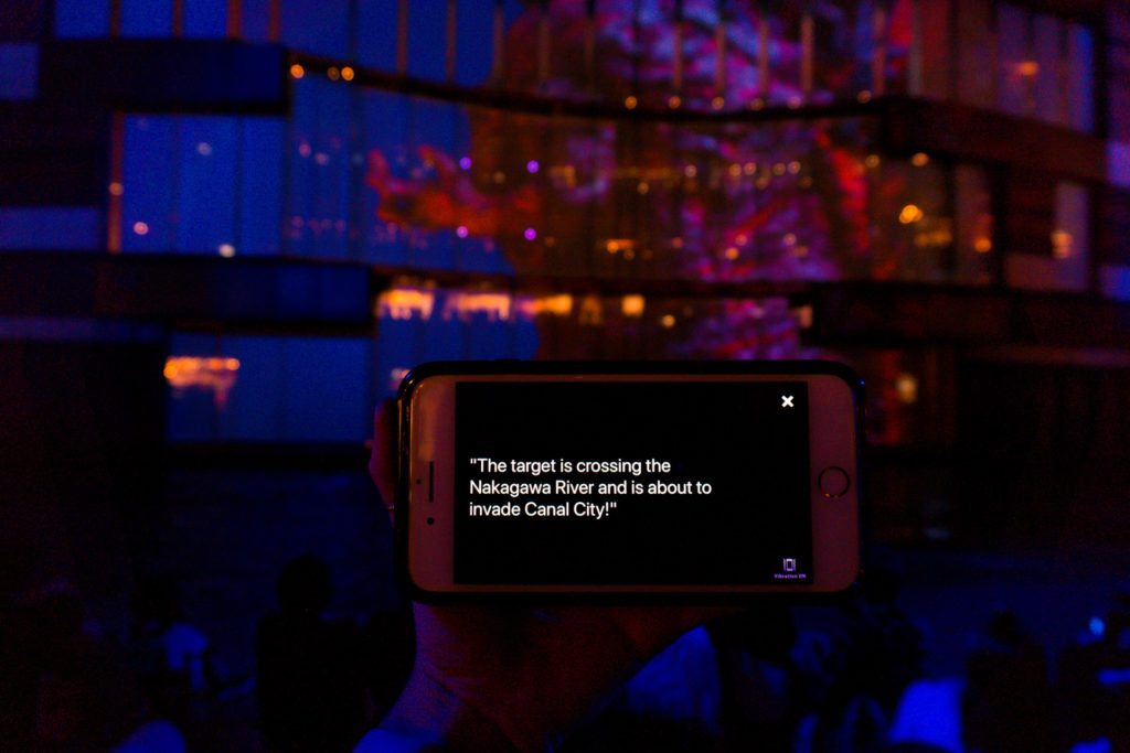 Hand holding a cell phone with text displayed that says "The target is crossing the Nakagawa River and is about to invade Canal City!"